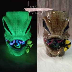 May include: A ceramic mug with a burlap texture and a green glow-in-the-dark design of a creature with a toothy grin and a yellow worm. The mug is a Halloween-themed design.