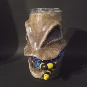 May include: A brown and beige ceramic tumbler with a burlap texture and a cartoon face. The face has black eyes and a blue and purple scarf. The tumbler has a yellow and black bee design on the front.