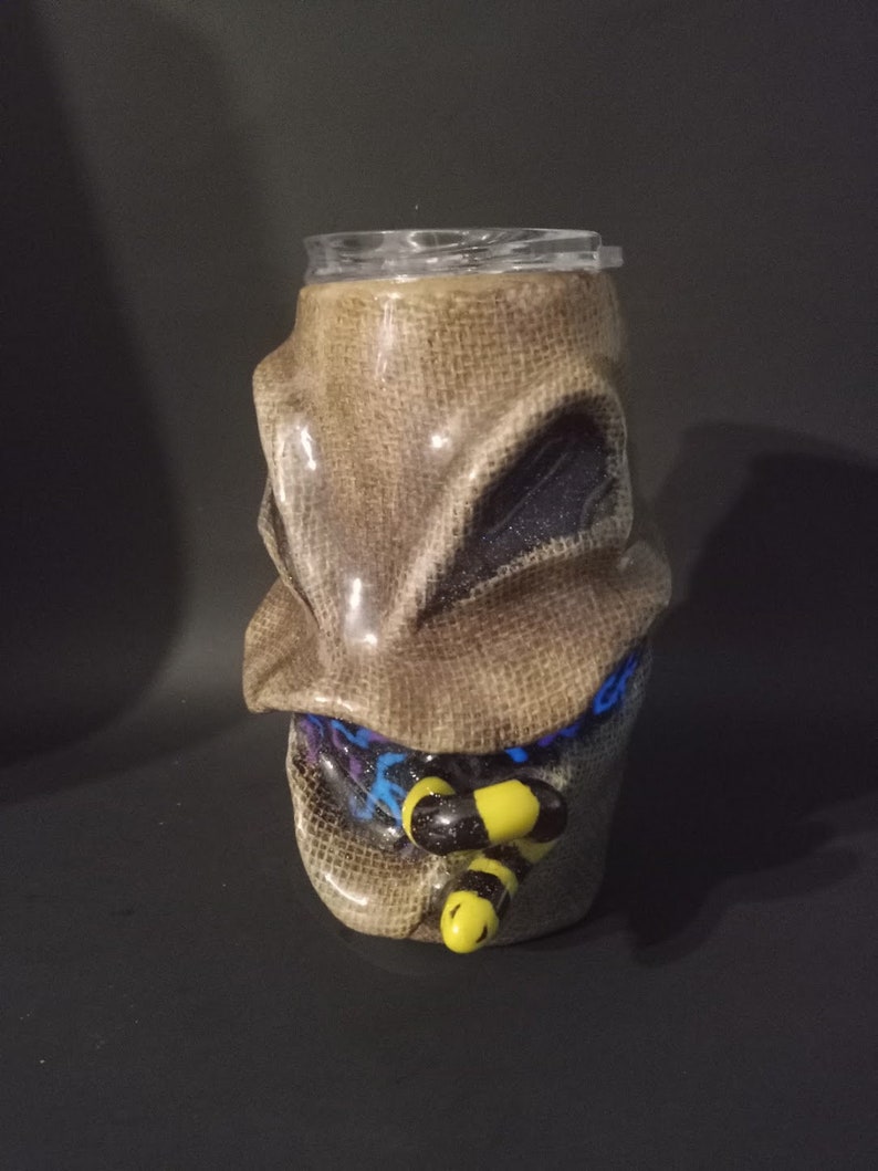 May include: A brown and beige ceramic tumbler with a burlap texture and a cartoon face. The face has black eyes and a blue and purple scarf. The tumbler has a yellow and black bee design on the front.