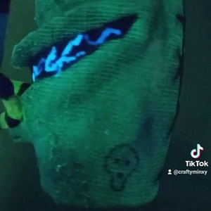 May include: A green fabric mask with a blue glow-in-the-dark design. The mask has a skull design on the front. The text "TikTok @craftyminxy" is visible in the bottom right corner.