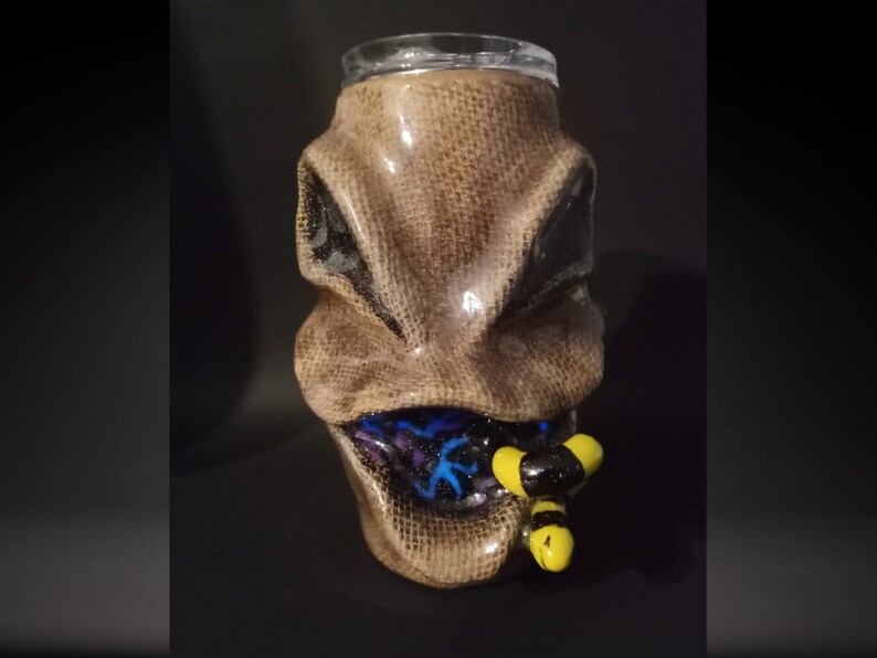 May include: A brown ceramic mug with a textured surface, resembling a face with a wide open mouth. The mug has a blue and purple design inside the mouth, and two yellow and black bees are sticking out of the mouth.