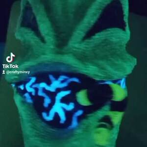 May include: A green glow-in-the-dark mask with black and blue details. The mask has a black eye mask with a blue and green pattern. The mask is made of a soft, fuzzy material.