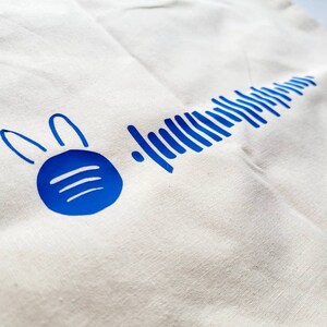 Può includere: Una borsa di tela bianca con un coniglio blu e un design a onde sonore blu. Il coniglio ha due orecchie e un corpo circolare con tre linee all'interno.