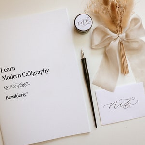 May include: A white booklet with the text "Learn Modern Calligraphy with Bewilderly" on the cover. A black calligraphy pen, a small jar of black ink, and a white card with the word "Nib" written in calligraphy are also pictured.
