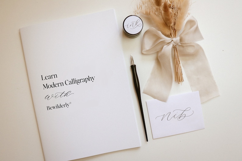 May include: A white booklet with the text "Learn Modern Calligraphy with Bewilderly" on the cover. A black calligraphy pen, a small jar of black ink, and a white card with the word "Nib" written in calligraphy are also pictured.