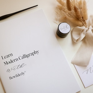 May include: A calligraphy kit with a white booklet that says "Learn Modern Calligraphy with Bewilderly", a black ink pot with a white label that says "ink", a white envelope with the word "Nib" written in calligraphy, a black calligraphy pen, and a bouquet of dried pampas grass tied with a white ribbon.