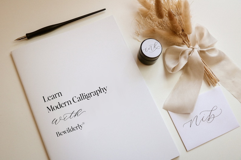 May include: A calligraphy kit with a white booklet that says "Learn Modern Calligraphy with Bewilderly", a black ink pot with a white label that says "ink", a white envelope with the word "Nib" written in calligraphy, a black calligraphy pen, and a bouquet of dried pampas grass tied with a white ribbon.
