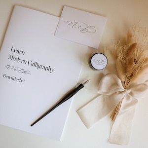 May include: A calligraphy kit with a white booklet that says "Learn Modern Calligraphy with Bewilderly", a black calligraphy pen, a small jar of ink, a white envelope with the word "Nib" written in calligraphy, and a bouquet of dried pampas grass tied with a white ribbon.