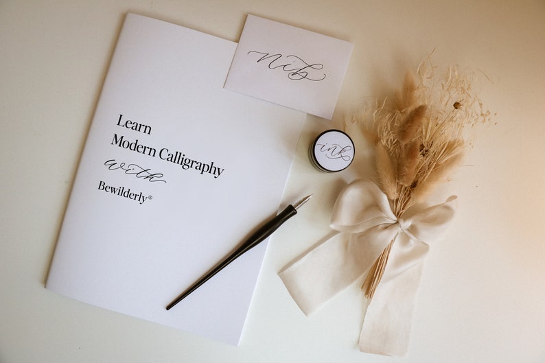 May include: A calligraphy kit with a white booklet that says "Learn Modern Calligraphy with Bewilderly", a black calligraphy pen, a small jar of ink, a white envelope with the word "Nib" written in calligraphy, and a bouquet of dried pampas grass tied with a white ribbon.