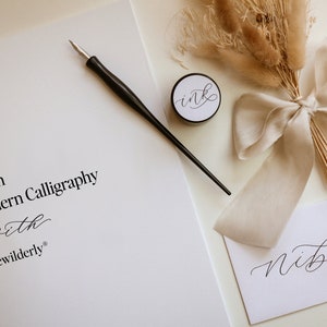 May include: Learn Modern Calligraphy with Bewilderly. A calligraphy kit with a black calligraphy pen, a small jar of ink, a white card with the word "nib" written in calligraphy, and a bouquet of dried pampas grass tied with a beige ribbon.