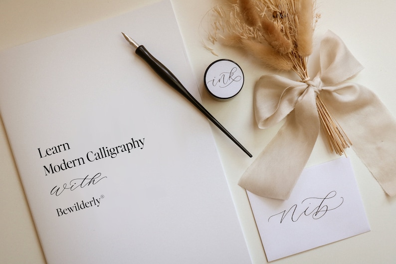 May include: Learn Modern Calligraphy with Bewilderly. A calligraphy kit with a black calligraphy pen, a small jar of ink, a white card with the word "nib" written in calligraphy, and a bouquet of dried pampas grass tied with a beige ribbon.
