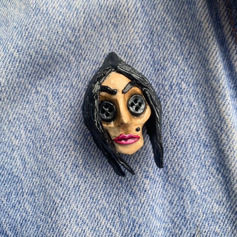 May include: A black and white ceramic pin of a woman with black hair and button eyes. The woman is wearing pink lipstick.