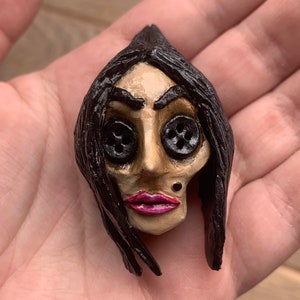 May include: A hand-painted ceramic doll head with black hair, button eyes, and a pink painted mouth. The doll's face is painted with a light tan color and has a small black dot on the chin.