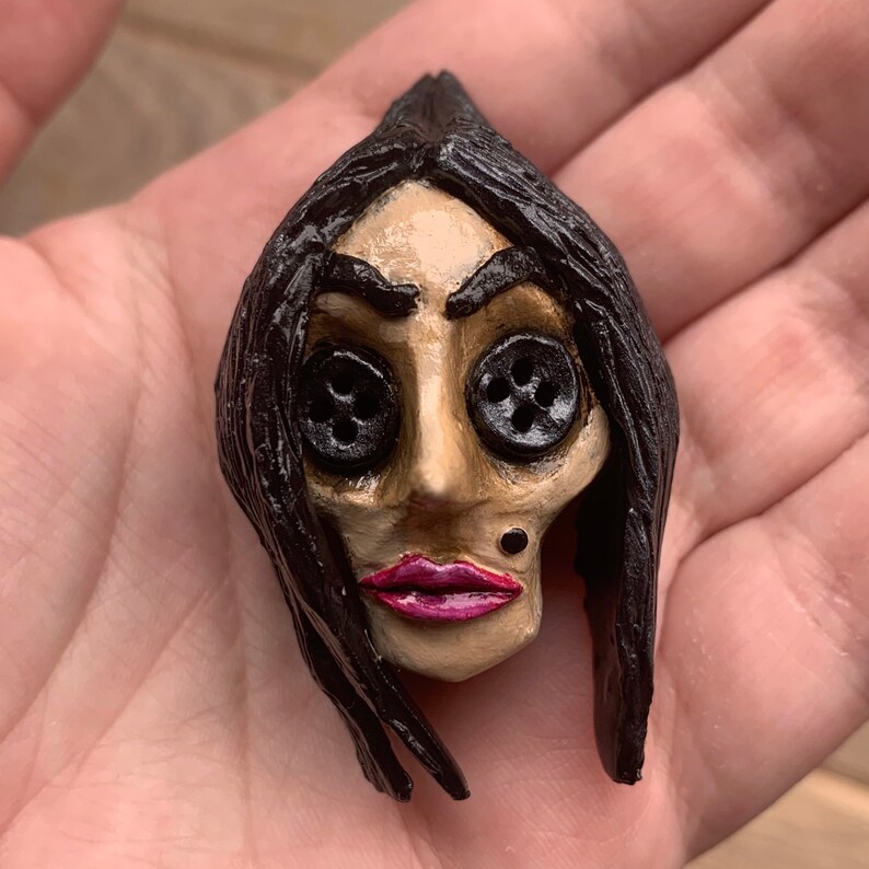 May include: A hand-painted ceramic doll head with black hair, button eyes, and a pink painted mouth. The doll's face is painted with a light tan color and has a small black dot on the chin.