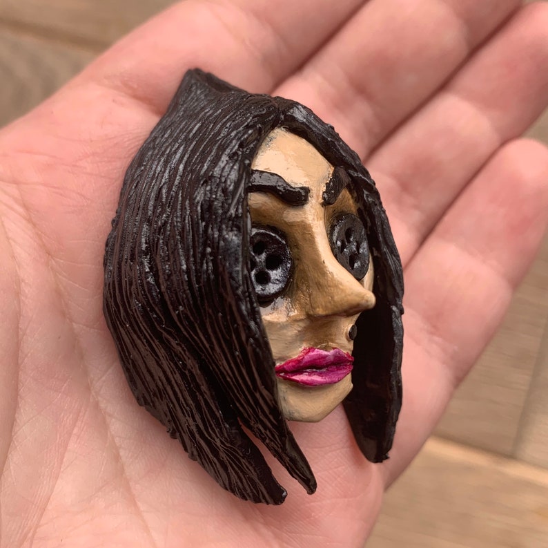 May include: A hand-painted ceramic figurine of a woman with black hair and button eyes. The figurine has a detailed face with a pink mouth and a realistic expression.