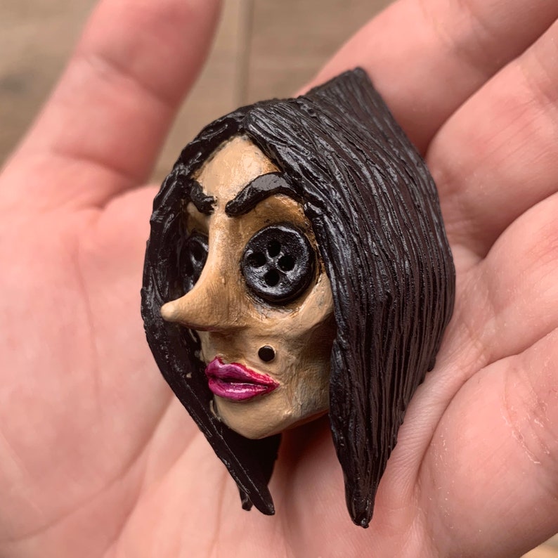 May include: A hand-painted ceramic doll head with black hair, button eyes, and a pink mouth. The doll has a sculpted nose and a small black dot on its chin.
