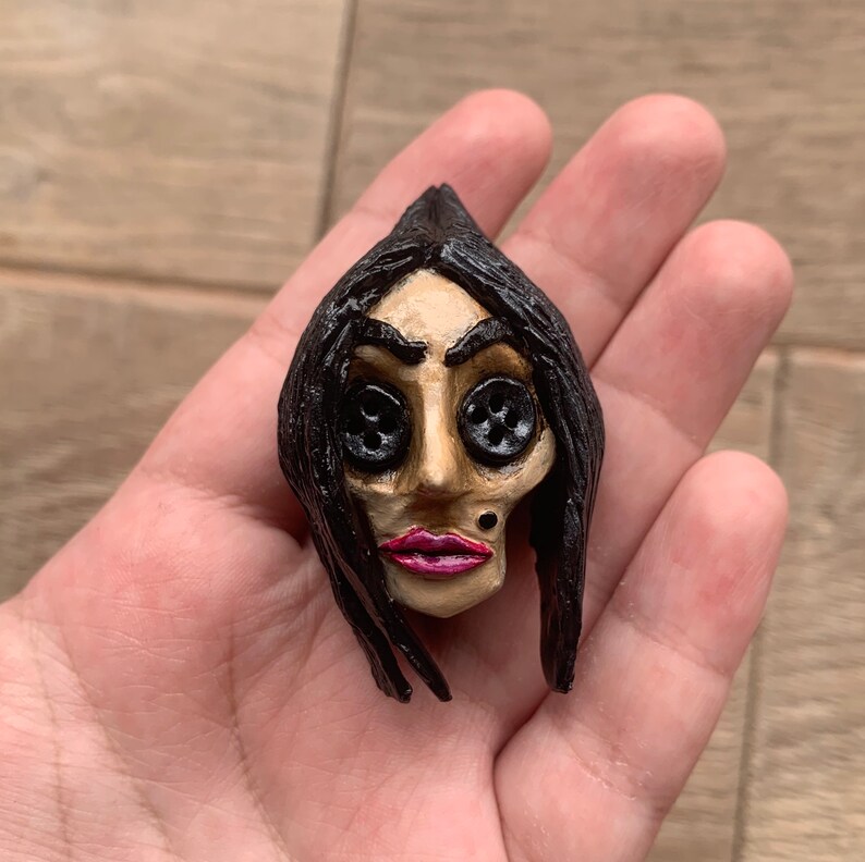 May include: A hand-crafted doll head with black hair, button eyes, and a painted face. The doll has a pink mouth and a small black dot on its chin.