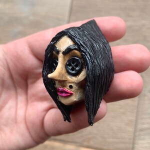 May include: A hand-crafted doll head with black hair, button eyes, and a pink painted mouth. The doll has a small black stud on its cheek.