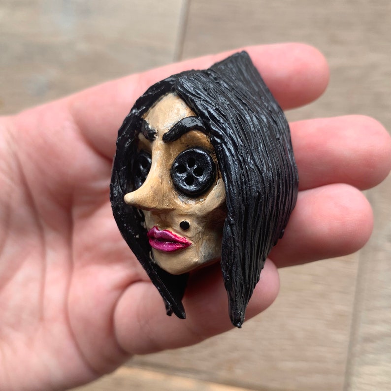 May include: A hand-crafted doll head with black hair, button eyes, and a pink painted mouth. The doll has a small black stud on its cheek.