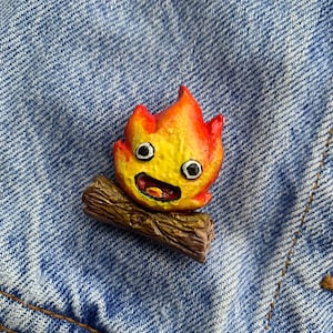 May include: A yellow and orange fire spirit with a smiling face, sitting on a brown log. The fire spirit has large eyes and a wide mouth.