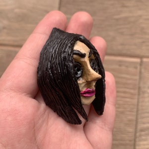 May include: A hand-painted figurine of a woman with black hair, brown eyes, and pink lips. The figurine has a detailed face and is made of clay.