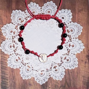 May include: Red macrame bracelet featuring black beads and a silver coin. The bracelet is displayed on a white lace doily, showcasing the intricate knotting and the contrast between the red cord, black beads, and silver coin.
