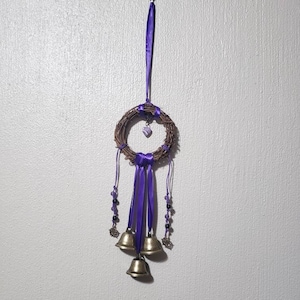 May include: A handcrafted wreath made of woven twigs, adorned with purple ribbon, a heart-shaped charm, and three small brass bells. The wreath is suspended by a purple ribbon, creating a decorative hanging ornament.