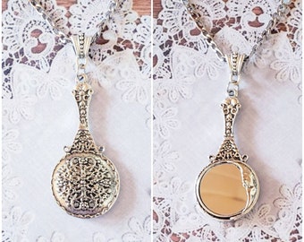 May include: A silver-toned pendant necklace featuring a handheld mirror design. The back of the circular pendant displays an intricate, raised filigree pattern, while the front reveals a smooth, reflective circular mirror surface attached to a chain.