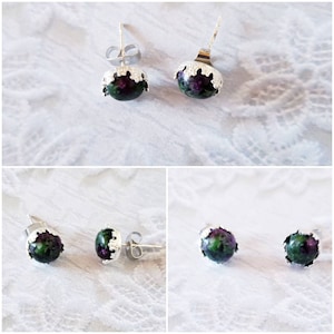 May include: A set of stud earrings featuring a dark green and purple stone, encased in a silver-colored metal setting. The earrings have a textured, decorative edge and a post-back closure. The image shows the earrings from multiple angles.