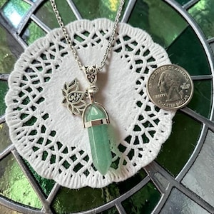 May include: A silver-toned necklace featuring a green crystal pendant and a lotus flower charm. The pendant is a light green, hexagonal shape. The necklace is displayed on a white, lacy doily, with a U.S. quarter dollar coin for size reference.