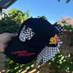 Puede incluir: Gorra de béisbol negra con un logotipo de la Ruta 66 y un diseño de bandera a cuadros. La gorra tiene el número 95 en llamas amarillas y rojas, y el nombre "Lightning McQueen" en escritura roja. Un lazo a cuadros está adjunto.