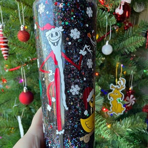 May include: A black glitter tumbler with a clear plastic lid. The tumbler features a white and red illustration of Jack Skellington dressed as Santa Claus. The tumbler also features a white snowflake design and a yellow duck toy.