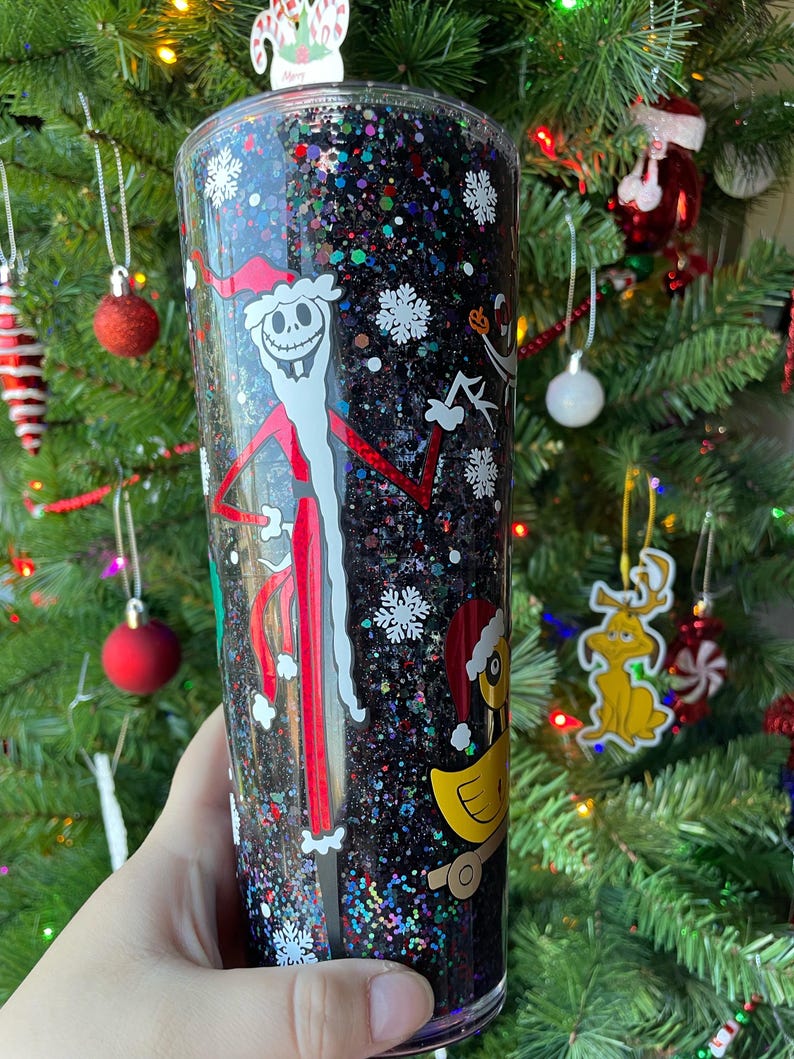 May include: A black glitter tumbler with a clear plastic lid. The tumbler features a white and red illustration of Jack Skellington dressed as Santa Claus. The tumbler also features a white snowflake design and a yellow duck toy.