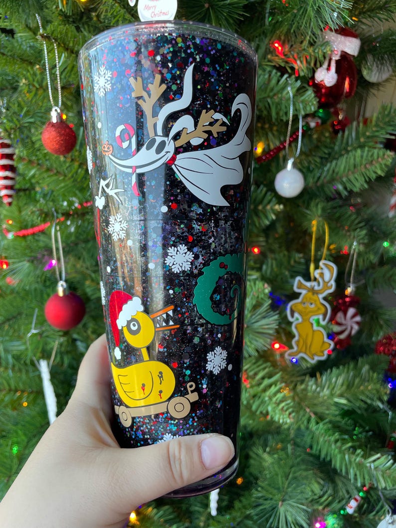 May include: A black glitter tumbler with a Christmas theme. The tumbler features a cartoon image of Jack Skellington from The Nightmare Before Christmas, a yellow duck with a Santa hat, and a green Grinch-like character. The tumbler is filled with colorful glitter.