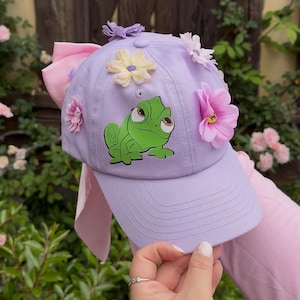 May include: A lavender baseball cap adorned with colorful fabric flowers and a green chameleon graphic. A large pink bow is attached to the back of the cap. The cap is held up by a person.