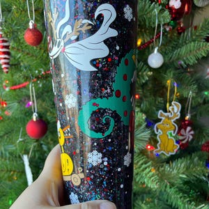 May include: A black glitter tumbler with a green, red, and white Christmas-themed design. The design features a creature from the movie "The Nightmare Before Christmas" and a Grinch-like character.