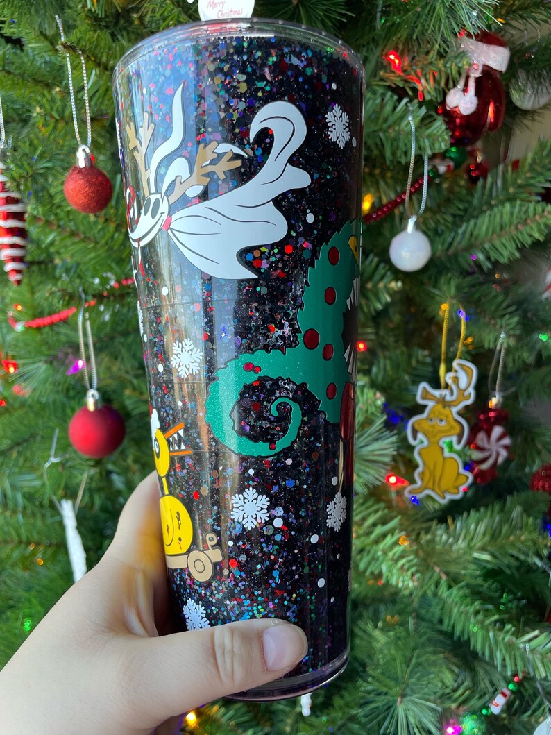 May include: A black glitter tumbler with a green, red, and white Christmas-themed design. The design features a creature from the movie "The Nightmare Before Christmas" and a Grinch-like character.