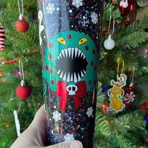 May include: A black glitter tumbler with a green Christmas wreath with a monster face and red bow. The wreath has a skull and crossbones on the bow. The tumbler is filled with black glitter and white snowflakes.