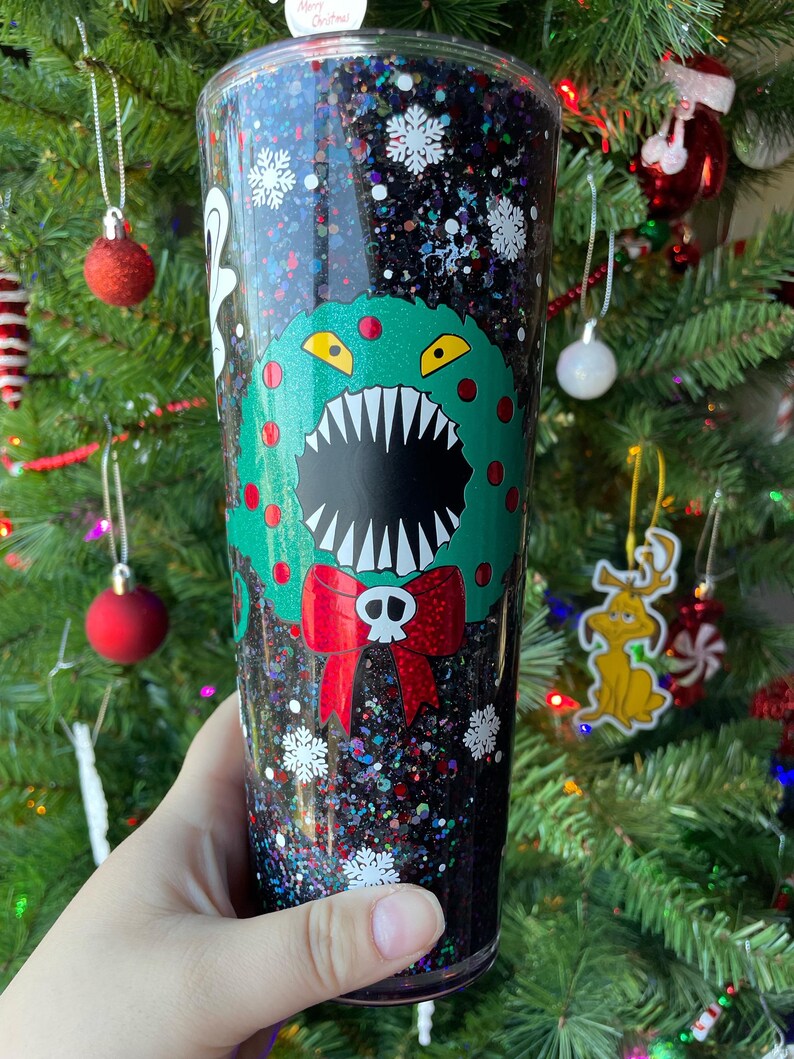 May include: A black glitter tumbler with a green Christmas wreath with a monster face and red bow. The wreath has a skull and crossbones on the bow. The tumbler is filled with black glitter and white snowflakes.