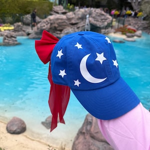 Peut inclure: Casquette de baseball bleue ornée d'étoiles blanches et d'un croissant de lune. Un grand nœud rouge est attaché à l'arrière de la casquette. La casquette est présentée devant un plan d'eau bleu.