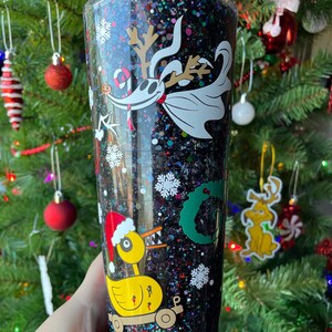 May include: A black glitter tumbler with a white and black image of Jack Skellington from The Nightmare Before Christmas. The tumbler also features a yellow duck with a red Santa hat and a green wreath.
