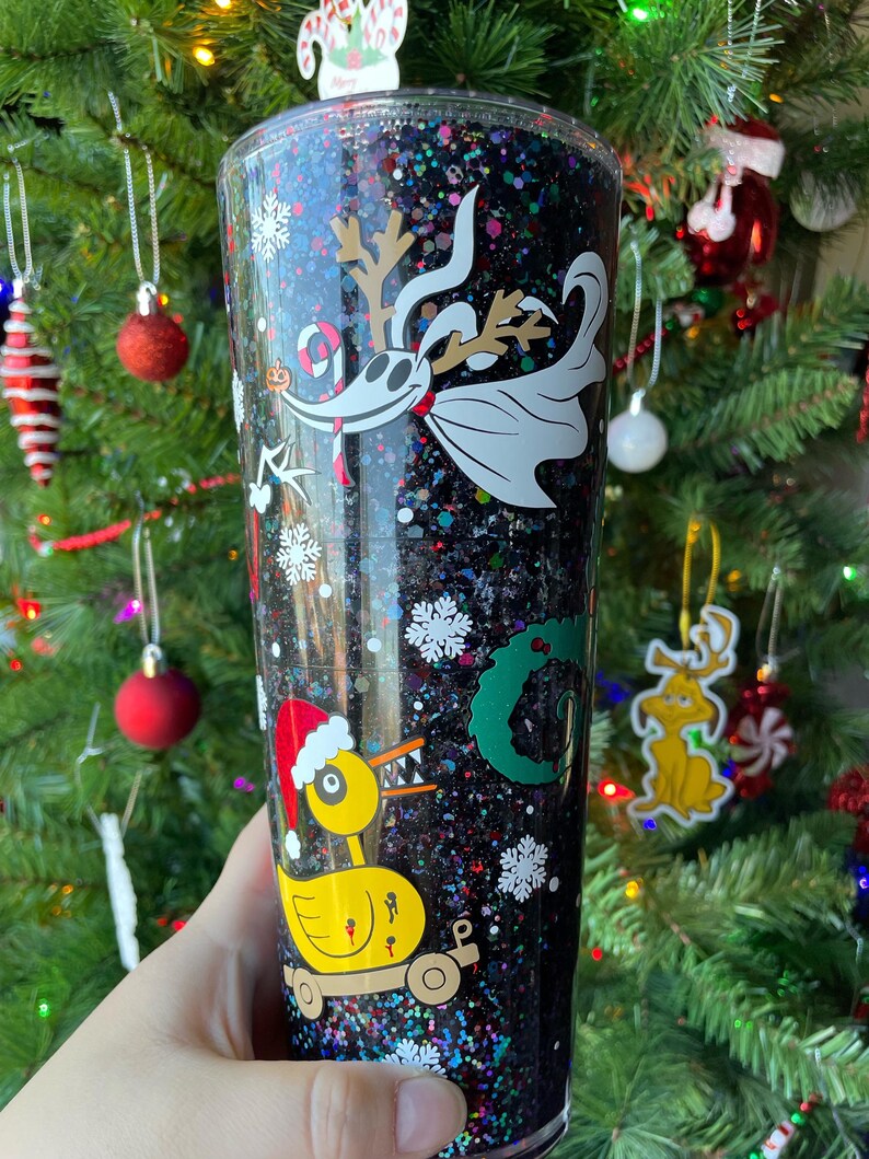 May include: A black glitter tumbler with a white and black image of Jack Skellington from The Nightmare Before Christmas. The tumbler also features a yellow duck with a red Santa hat and a green wreath.