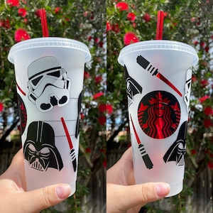May include: Two clear plastic cups with lids and red straws. The cups are decorated with Star Wars characters, including Darth Vader, Stormtroopers, and Yoda. The cups are perfect for enjoying your favorite beverage.