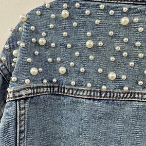 Może przedstawiać: Zbliżenie jasnoniebieskiej kurtki dżinsowej ozdobionej licznymi sztucznymi perłami o r&oacute;żnych rozmiarach. Perły są rozrzucone na plecach i ramionach kurtki, dodając odrobinę elegancji do tego codziennego elementu garderoby.