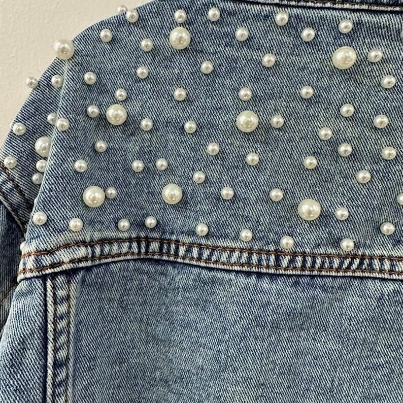 Może przedstawiać: Zbliżenie jasnoniebieskiej kurtki dżinsowej ozdobionej licznymi sztucznymi perłami o r&oacute;żnych rozmiarach. Perły są rozrzucone na plecach i ramionach kurtki, dodając odrobinę elegancji do tego codziennego elementu garderoby.