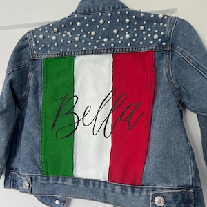 Może przedstawiać: Jeansowa kurtka z namalowaną na plecach włoską flagą. Flaga ma zielone, białe i czerwone pionowe paski, z napisem "Bella" w kursywie. Ramiona kurtki zdobią małe, białe perły.