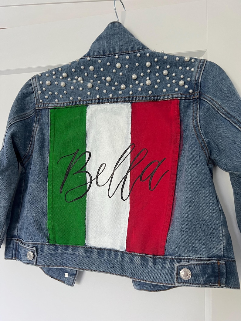 Może przedstawiać: Jeansowa kurtka z namalowaną na plecach włoską flagą. Flaga ma zielone, białe i czerwone pionowe paski, z napisem "Bella" w kursywie. Ramiona kurtki zdobią małe, białe perły.