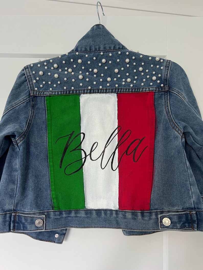 Może przedstawiać: Jeansowa kurtka z namalowaną na plecach włoską flagą. Flaga ma zielone, białe i czerwone paski, ze słowem "Bella" w czarnym skrypcie. Kołnierz jest ozdobiony małymi, białymi perłami.