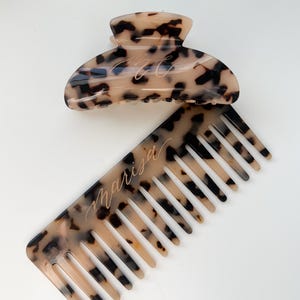 Könnte beinhalten: Ein Kamm und eine Haarklammer aus Schildpatt mit dem eingravierten Namen "Marisa" auf dem Kamm.