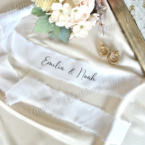 May include: White silk ribbon with frayed edges, featuring the names "Emilia & Noah" in elegant black script. The ribbon is draped on a satin surface, accompanied by gold earrings and floral accents, creating a romantic wedding theme.